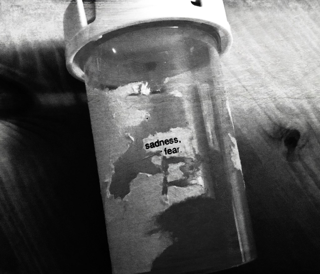 Black and white close up of a standard USA medication bottle resting on a wooden table (grain visible). The label has been scratched off except for “sadness, fear.”  Hight contrast, some distress. 